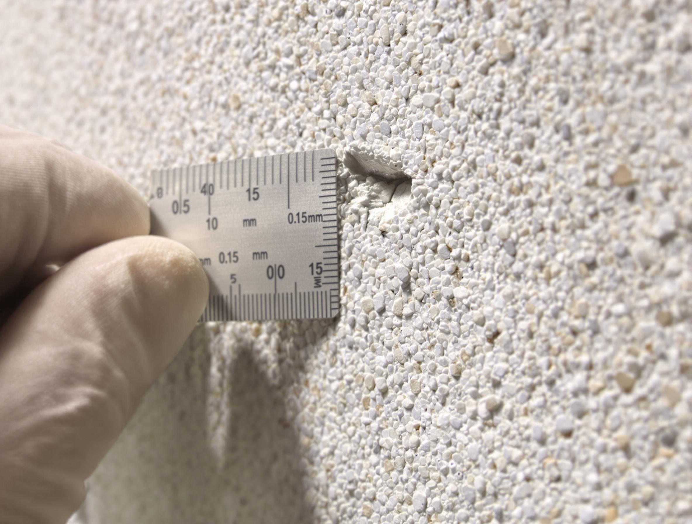 Gros plan d’une microfissure de 0,15 mm sur un enduit à la chaux éclairage rasant texture granitée mortier règle graduée main gantée.
