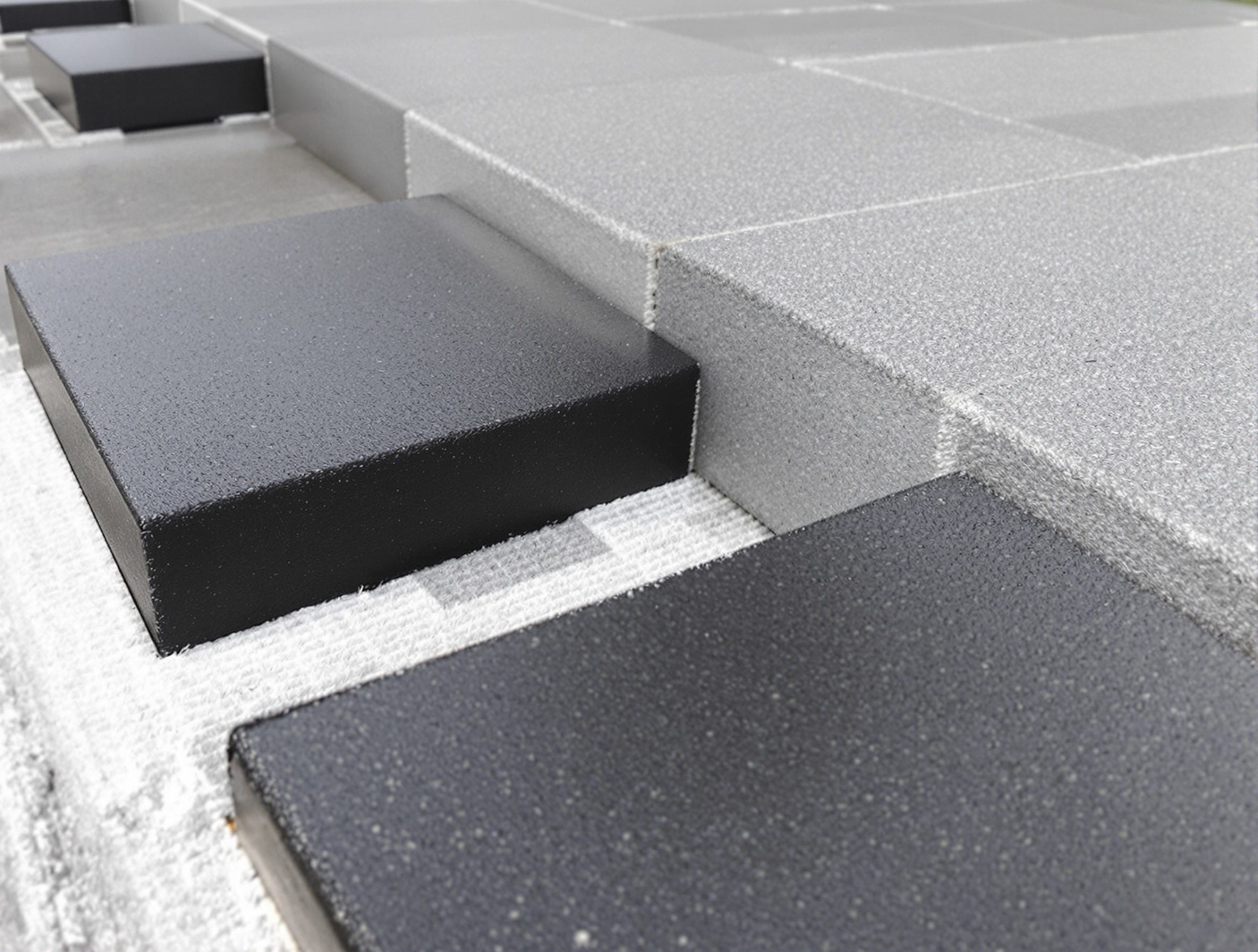 Cales amortisseurs noires posées sur têtes de plots terrasse, joints ouverts visibles et film géotextile sous-jacent.